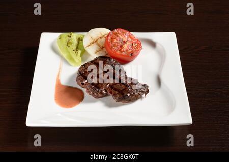 Geröstetes Fleisch von Rindersteaks aus Marmor mit gegrilltem Gemüse aus Tomaten, Zwiebeln und Paprika auf einem quadratischen Teller auf einem Holztisch. Fotos für das Restaurant Stockfoto