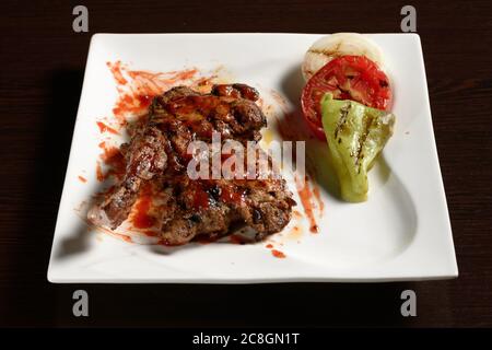 Gegrillte Hähnchenschenkel mit gegrilltem Gemüse aus Tomaten, Zwiebeln und Paprika auf einem quadratischen Teller auf einem Holztisch. Fotos für Restaurant und Cafe Me Stockfoto