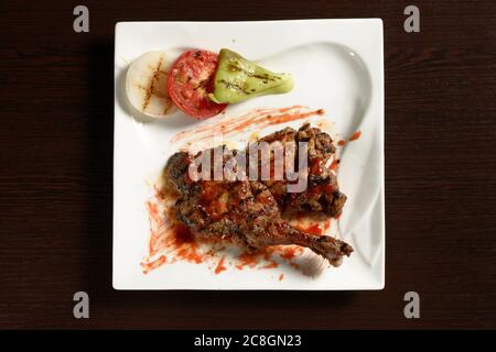 Barbecue Chicken Oberschenkel mit gegrilltem Gemüse von Tomaten, Zwiebeln und Paprika auf einem quadratischen Teller auf einem Holztisch. Draufsicht. Fotos für Restaurant A Stockfoto