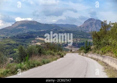 Abstieg auf einer asphaltierten Straße, mit Feldern und Bergen im Hintergrund, Stockfoto