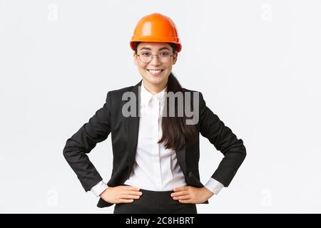 Professionelle lächelnde asiatische Bauingenieurin, Architekt in schwarzem Anzug und Sicherheitshelm, die bei der Arbeit zufrieden, zufrieden stehend Stockfoto