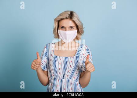 Junge kaukasische Frau trägt eine Anti-Grippe-Maske und Kleid, während das ähnliche Zeichen auf einer blauen Studiowand gestikulieren Stockfoto