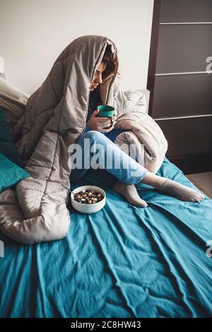 Charmante Dame mit lockigem Haar, Milch trinken und Getreide essen im Bett mit einer Bettdecke bedeckt Stockfoto
