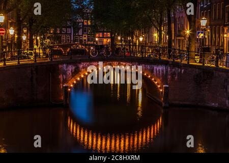 Amsterdams historische Reihenhäuser leuchten nachts entlang der ruhigen Kanäle und ihre Reflexionen schimmern über die ruhige Uferpromenade. Stockfoto