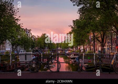 Amsterdams historische Reihenhäuser leuchten nachts entlang der ruhigen Kanäle und ihre Reflexionen schimmern über die ruhige Uferpromenade. Stockfoto