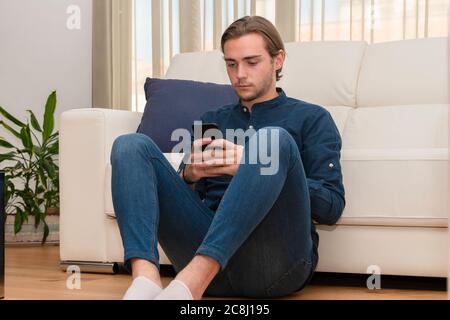 Hübscher junger Mann, der auf dem Boden sitzt und sich auf ein Sofa stützt, das auf sein Telefon auf einem unfokussierten Hintergrund fokussiert ist. Kommunikations- und Freizeitkonzept. Stockfoto