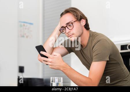 Schöner junger Mann trägt eine Brille, die sein Haar fixiert, während er sein Telefon auf einem unscharf Hintergrund verwendet. Kommunikations- und Freizeitkonzept. Stockfoto