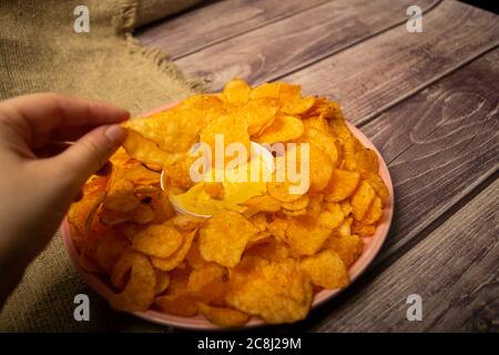 Das Mädchen nimmt einen Chip aus einem runden Gericht mit Kartoffelchips und einem Topf mit Käsesauce in der Mitte des Tellers. Nahaufnahme Stockfoto