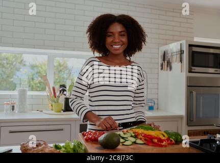 Fröhliche junge Frau mit lockigen Haaren hacken Gemüse für Salat in der modernen Küche Stockfoto