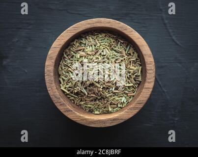 Getrockneter Rosmarin - Salvia rosmarinus in Holzschüssel. Kraut mit evegrünen nadelartigen Blättern. Aufnahme über dem Kopf mit dunklem Hintergrund. Stockfoto