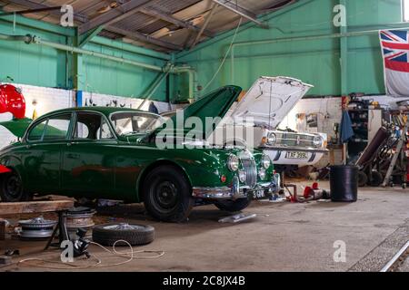 Jaguar Mk2 Limousine und Ford Cortina stehen mit ihren Motorhauben in einer Oldtimer-Restaurierungswerkstatt, mit Reifen, Rädern und Werkzeugen um sie herum Stockfoto