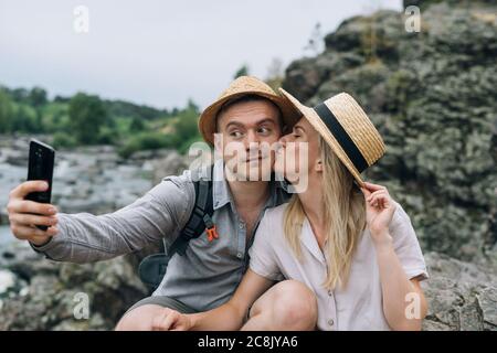 Glückliche junge Paar Reisende in lässigen Outfits nehmen Selfie auf Handy am Berg Fluss Hintergrund. Lokaler Tourismus, Wochenend-Trip-Konzept Stockfoto