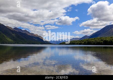 Das ruhige Wasser des South Mavora Lake mit bewaldeten Bergen auf jeder Seite und Bergen in der Ferne. Weiße flauschige Wolken an einem blauen Himmel. Stockfoto