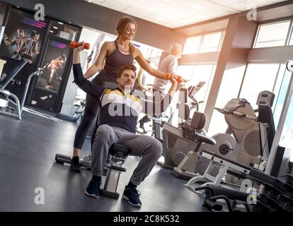 Junge weibliche Trainerin hilft dem jungen Mann, die Hanteln zu heben Im Fitnessstudio Stockfoto