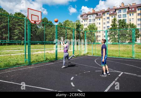 Junger Mann in blauem T-Shirt und Denim-Shorts, der am sonnigen Sommertag mit seiner Mutter auf dem Sportplatz Basketball spielt Stockfoto