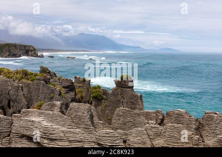 Die berühmten Pancake Rocks auf Dolomitenspitze mit Blick über das Meer auf die von Wolken bedeckten Berge. Dolomit Point, Südinsel, Neuseeland Stockfoto