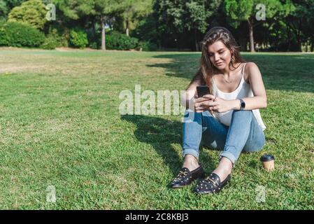 Schöne junge kaukasische Frau sitzt auf dem Gras eines städtischen Parks SMS mit einem Smartphone Stockfoto