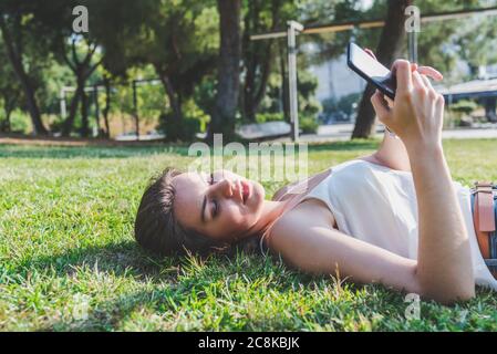 Schöne junge kaukasische Frau, die auf dem Gras eines Stadtparks liegt und ein Telefon benutzt Stockfoto