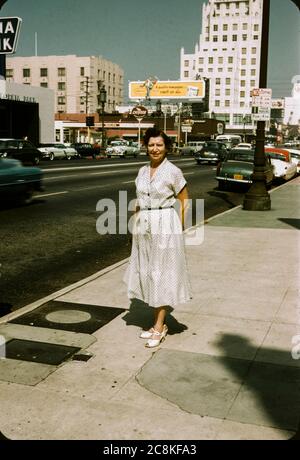Frau, die in den 1950er Jahren auf dem Bürgersteig in der Innenstadt von Los Angeles, Kalifornien, stand. Plakate und 50er Jahre Autos sind im Hintergrund, einschließlich einer Plakatwand für die Los Angeles Times Zeitung. Stockfoto