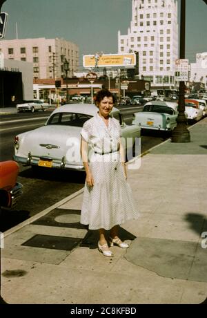 Frau, die 1957 auf dem Bürgersteig in der Innenstadt von Los Angeles, Kalifornien, stand. Plakate und 50er Jahre Autos sind im Hintergrund, einschließlich einer Plakatwand für die Los Angeles Times Zeitung. Los Angeles Street Scene 1957 Kodachrome Red Mount Cars Stockfoto