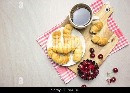 Frühstück mit hausgemachten Croissants, Kaffee und Süßkirschen auf hellem texturierten Hintergrund, Draufsicht mit Kopierraum. Stockfoto