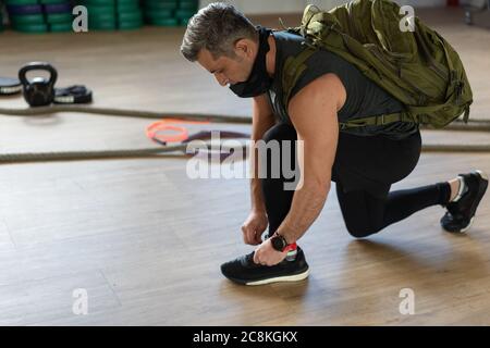 Motivierter Instruktor kniet sich nieder und bindet seine Schuhschnur mit Rucksack und Trainingsmaske. Beginn der Schulung. Für Boot Camp und Fitness-Konzept. Stockfoto