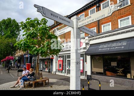 Ein lokales Schild in Dulwich Village mit Menschen und Geschäften. Das Schild zeigt Ihnen die Wegbeschreibung zu den Sehenswürdigkeiten von Dulwich und den nahegelegenen Orten. Dulwich liegt im Süden Londons Stockfoto