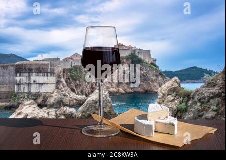 Glas Rotwein mit Brie-Käse mit Blick auf die Festungsmauern, felsige Küste und blaues Meer, Dubrovnik, Kroatien. Atemberaubender Blick auf die Stadtmauer von Dubrovnik Stockfoto