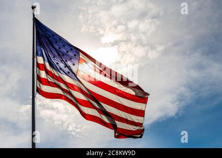 Große schöne amerikanische Flagge leuchtet von der Hintergrundbeleuchtung von der Sonne, gegen bewölkten Himmel Stockfoto