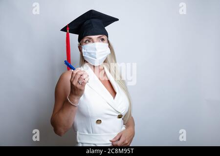 Eine schöne Absolventin, die eine medizinische Schutzmaske auf ihrem Gesicht und eine Abschlusskappe auf ihrem Kopf trägt, hält einen Stift in ihrer Hand. Hochwertige Fotos Stockfoto