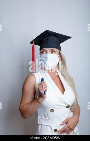 Eine schöne Absolventin, die eine medizinische Schutzmaske auf ihrem Gesicht und eine Abschlusskappe auf ihrem Kopf trägt, hält einen Stift in ihrer Hand. Hochwertige Fotos Stockfoto
