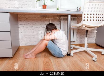 Trauriges und unglückliches Kind, das auf dem Boden unter dem Tisch sitzt. Verärgert, Junge Stockfoto