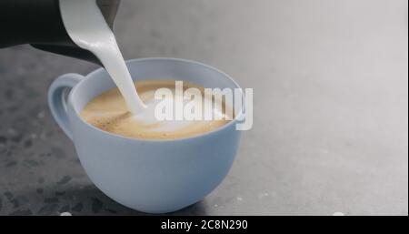 Gieße gedämpfte Milch in Espresso, um Cappuccino in Blau zuzubereiten becher auf betonem Hintergrund Stockfoto