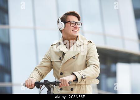 Grafik Taille up Porträt von fröhlichen jungen Geschäftsmann tragen Trenchcoat und Kopfhörer wegschauen, während Reiten Elektroroller mit Glas Stadt bui Stockfoto