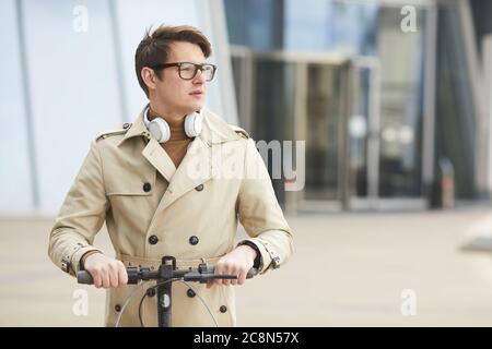 Taille up Porträt des modernen jungen Geschäftsmann Reiten Elektroroller in Richtung Kamera und Blick weg mit städtischen Gebäuden im Hintergrund, Kopie Spa Stockfoto