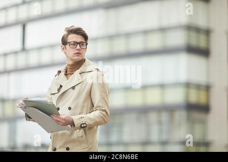 Waist up Porträt des jungen Geschäftsmann tragen Trenchcoat und halten Zwischenablage, während im Freien stehen und wegschauen in städtischen Umgebung, Kopie s Stockfoto