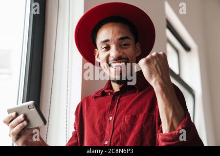 Bild eines schönen afrikanischen optimistischen jungen Mann posiert drinnen in der Nähe Fenster mit Handy, während macht Sieger Geste Stockfoto