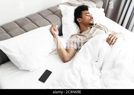 Foto von jungen afroamerikanischen Mann mit Schnurrbart schlafen in weißem Bett in hellen Raum Stockfoto