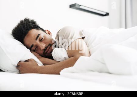 Foto von jungen afroamerikanischen Mann mit Schnurrbart schlafen in weißem Bett in hellen Raum Stockfoto