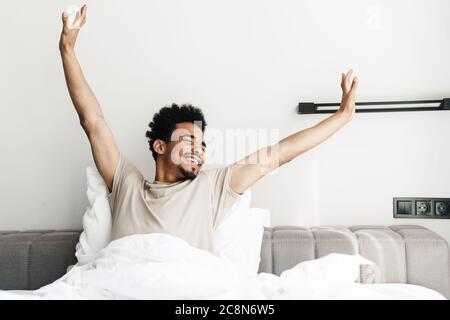 Foto von zufrieden african american Mann mit Schnurrbart Stretching seinen Körper im Bett in hellen Raum Stockfoto