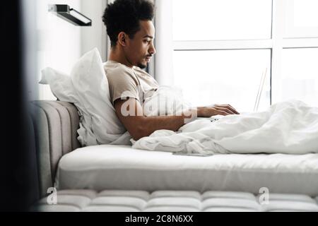 Foto eines konzentrierten afroamerikanischen Mannes, der mit einem Laptop arbeitet, während er im Bett im hellen Zimmer liegt Stockfoto