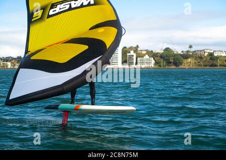 Ein junger Mann wingfoils im Hafen von Auckland, mit einem handaufblasbaren Flügel und Reiten ein Tragflächenboot Surfbrett. Grünes Meer und wolkig blauer Himmel. Stockfoto