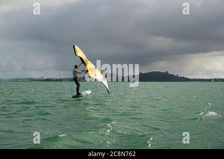 Ein junger Mann wingfoils im Hafen von Auckland, mit einem handaufblasbaren Flügel und Reiten ein Tragflächenboot Surfbrett. Grünes Meer und wolkig blauer Himmel. Stockfoto