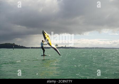 Ein junger Mann wingfoils im Hafen von Auckland, mit einem handaufblasbaren Flügel und Reiten ein Tragflächenboot Surfbrett. Grünes Meer und wolkig blauer Himmel. Stockfoto