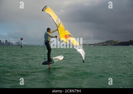 Ein junger Mann wingfoils im Hafen von Auckland, mit einem handaufblasbaren Flügel und Reiten ein Tragflächenboot Surfbrett. Grünes Meer und wolkig blauer Himmel. Stockfoto