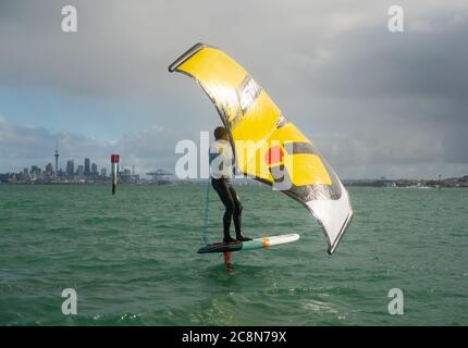 Ein junger Mann wingfoils im Hafen von Auckland, mit einem handaufblasbaren Flügel und Reiten ein Tragflächenboot Surfbrett. Grünes Meer und wolkig blauer Himmel. Stockfoto