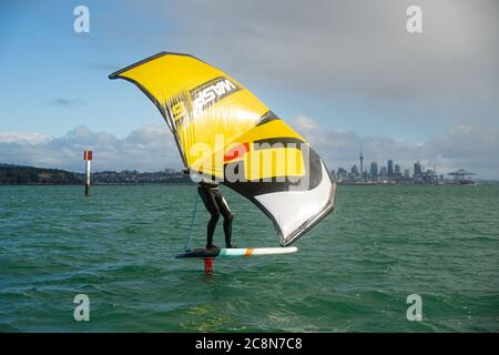 Ein junger Mann wingfoils im Hafen von Auckland, mit einem handaufblasbaren Flügel und Reiten ein Tragflächenboot Surfbrett. Grünes Meer und wolkig blauer Himmel. Stockfoto