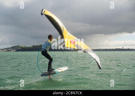 Ein junger Mann wingfoils im Hafen von Auckland, mit einem handaufblasbaren Flügel und Reiten ein Tragflächenboot Surfbrett. Grünes Meer und wolkig blauer Himmel. Stockfoto