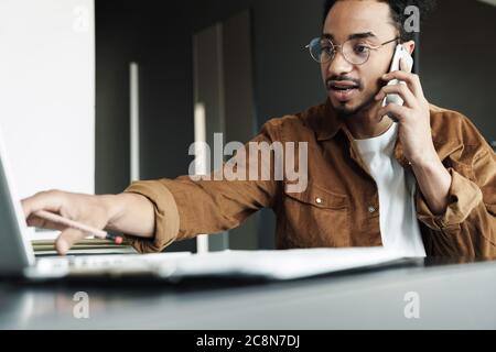 Foto von fokussierten afroamerikanischen Mann auf Handy sprechen, während mit Laptop im Wohnzimmer arbeiten Stockfoto