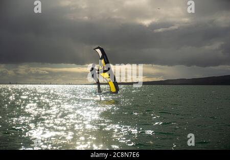 Ein junger Mann wingfoils im Hafen von Auckland, mit einem handaufblasbaren Flügel und Reiten ein Tragflächenboot Surfbrett. Grünes Meer und wolkig blauer Himmel. Stockfoto